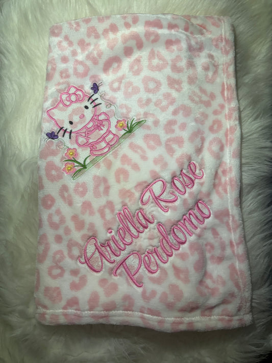 Pink leopard print blanket with Hello Kitty and embroidered name on a fluffy white surface